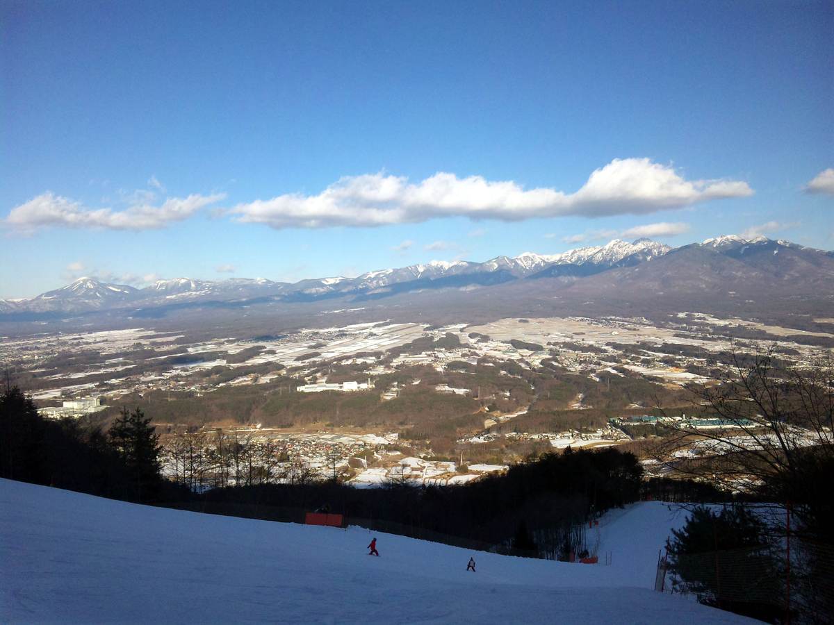 Two snowboarders descend a piste at Fujimi Panorama with a wide view over the Yatsugatake mountain range and the valley below