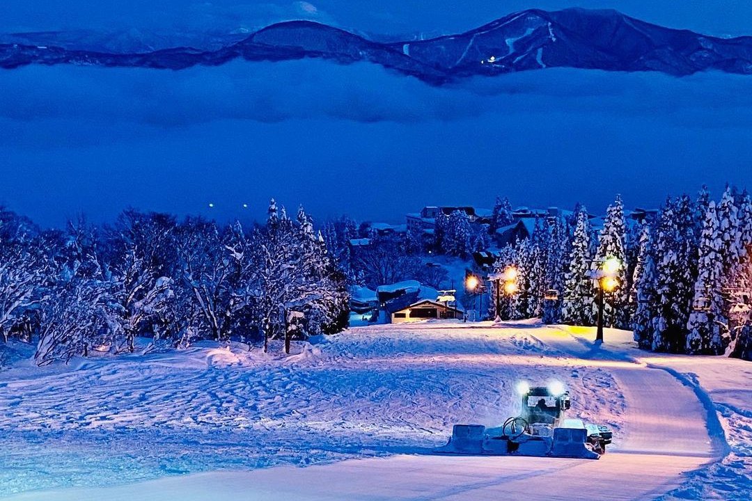 A piste groomer on a groomed run at dusk, with snow-covered trees, lit buildings and a valley of cloud below a ridge